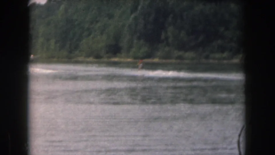 1961 Michigan home-movie frame showing a distant water skier moving across a lake with trees along the far shore.