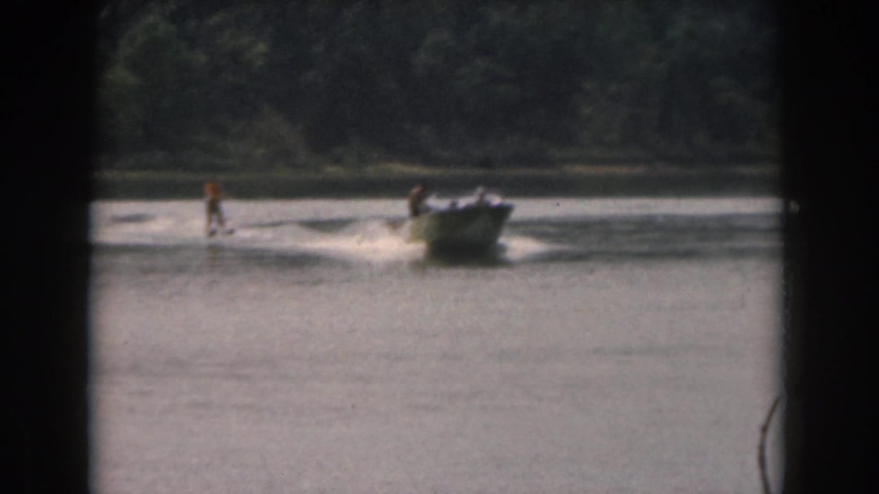 1961 Grand Rapids area lake scene with a small motorboat pulling a distant water skier past a wooded shoreline.