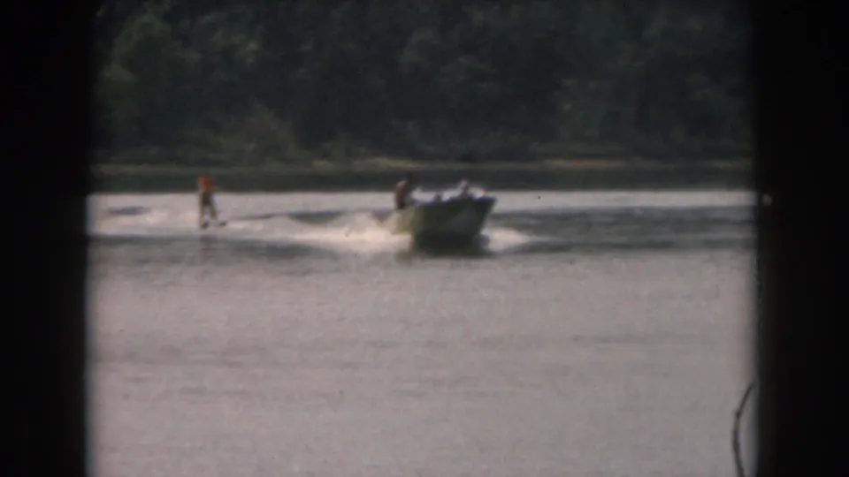 1961 Grand Rapids area lake scene with a small motorboat pulling a distant water skier past a wooded shoreline.