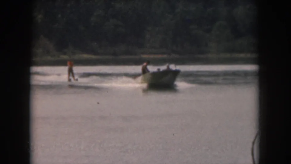 Vintage 8mm frame of a water skier following a small motorboat across a Michigan lake, with trees in the background.