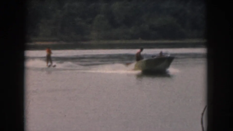 1961 Grand Rapids-area home-movie frame showing a distant water skier, tow boat, wake, and wooded lake shore.