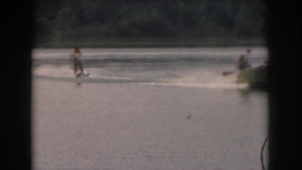 Home-movie view of a skier in an orange life vest crossing open lake water near a partially visible tow boat.