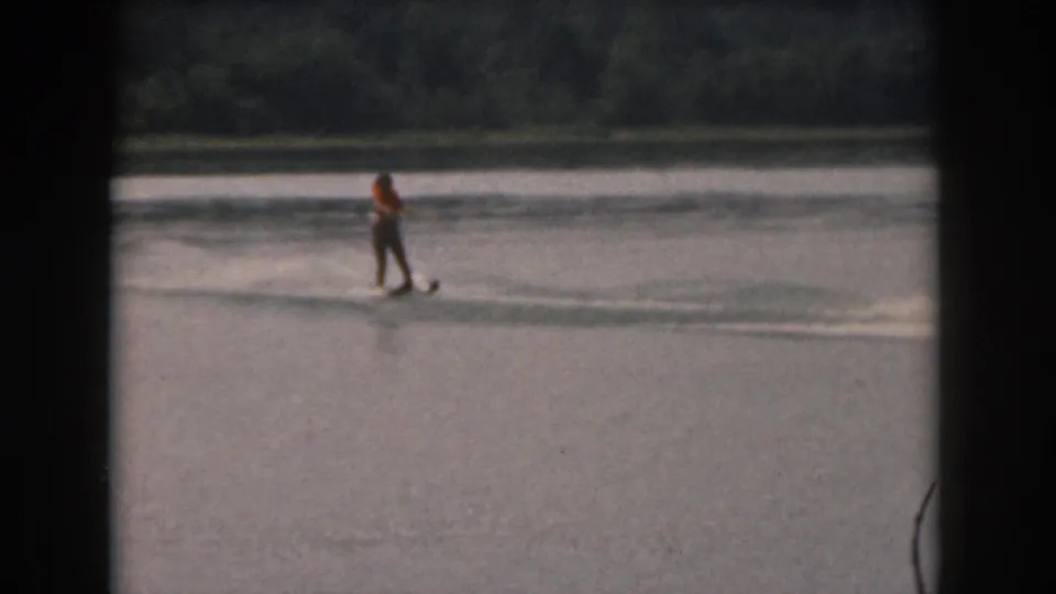 Distant 8mm frame of a water skier wearing an orange vest, balanced on skis over calm water in Michigan footage.