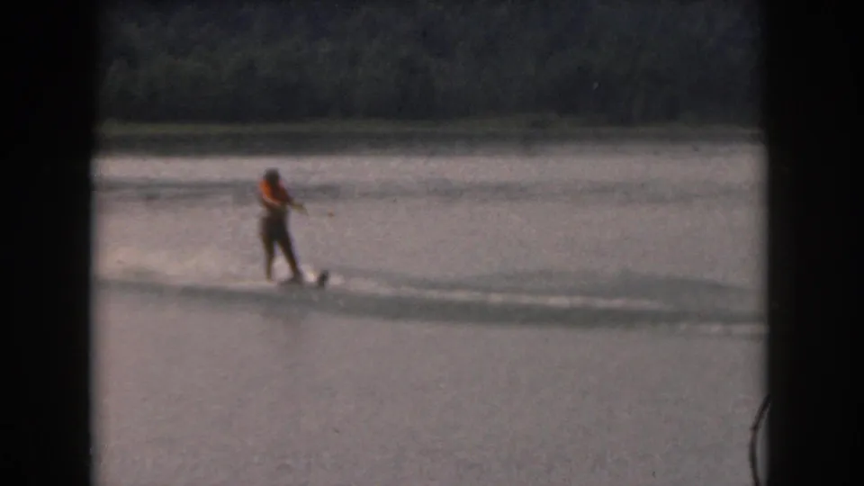 1961 home-movie still of a distant water skier on a tree-lined lake, framed by dark 8mm film edges.