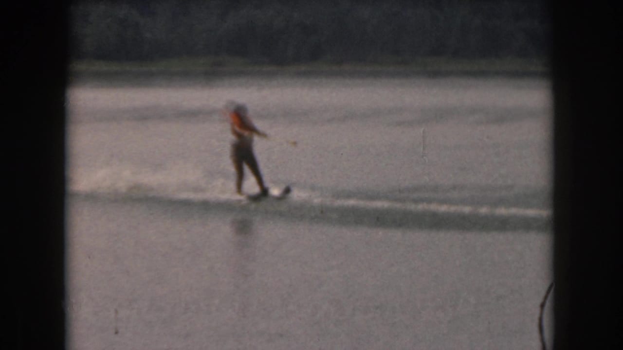 Distant woman water skiing across calm water inside a dark 8mm film frame.
