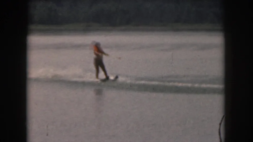 Distant 1961 Grand Rapids home-movie frame of a woman in an orange life jacket water skiing across a lake.