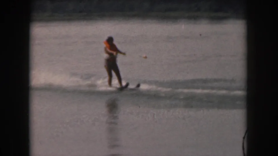 Woman in an orange life jacket holds a tow rope while water skiing on a calm Michigan lake in 1961 footage.