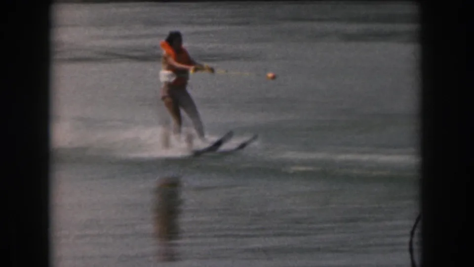 Closer 8mm frame of the woman water skiing in an orange life jacket, with yellow tow rope visible over the lake.