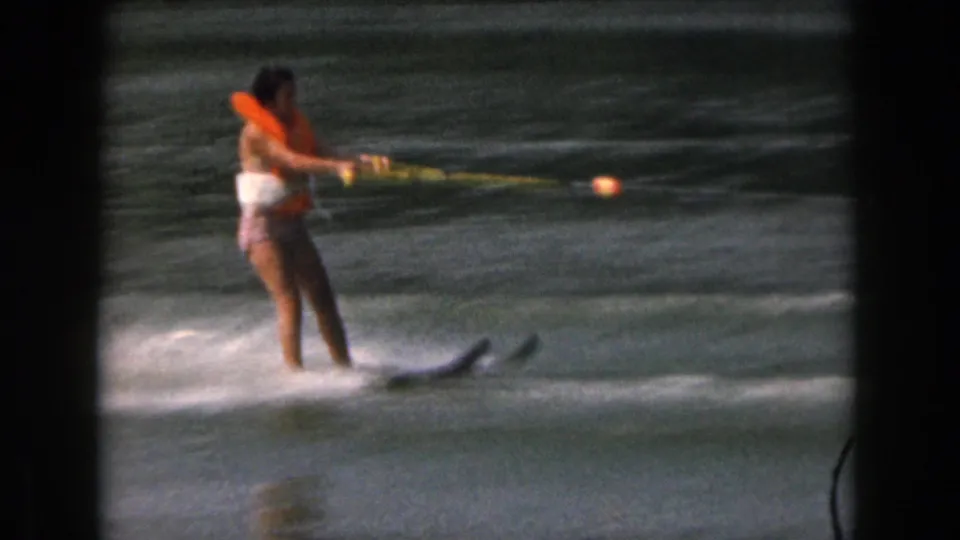 1961 home-movie frame showing a woman in a bright life jacket water skiing, with the tow rope extending across the lake.