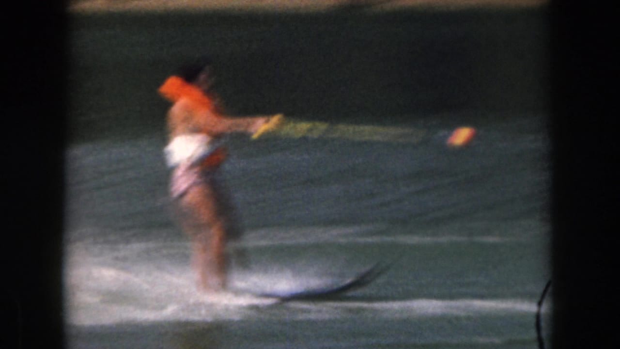 1961 Michigan home-movie frame of a woman on two water skis, wearing an orange life jacket as she moves across the lake.