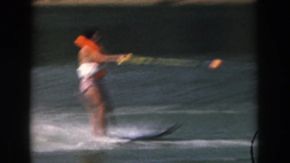 1961 Michigan home-movie frame of a woman on two water skis, wearing an orange life jacket as she moves across the lake.