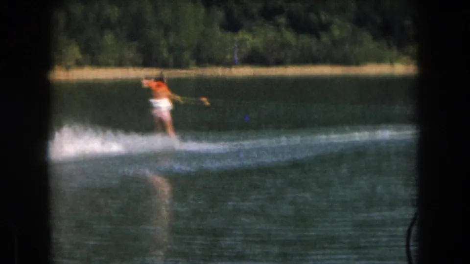 1961 Grand Rapids-area lake scene showing a woman water skiing at a distance with trees and shoreline behind her.