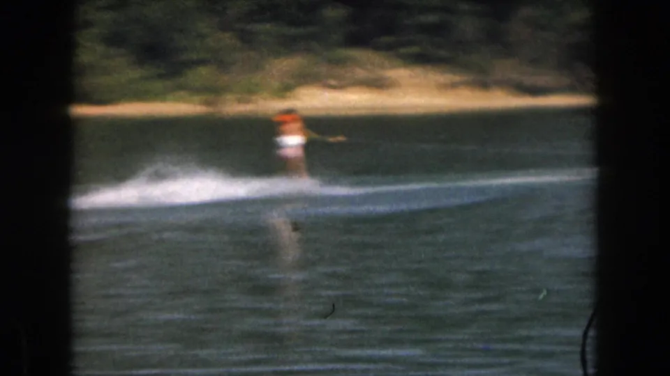 1961 home-movie still of a woman water skiing across a Michigan lake, framed by dark 8mm borders and shoreline trees.
