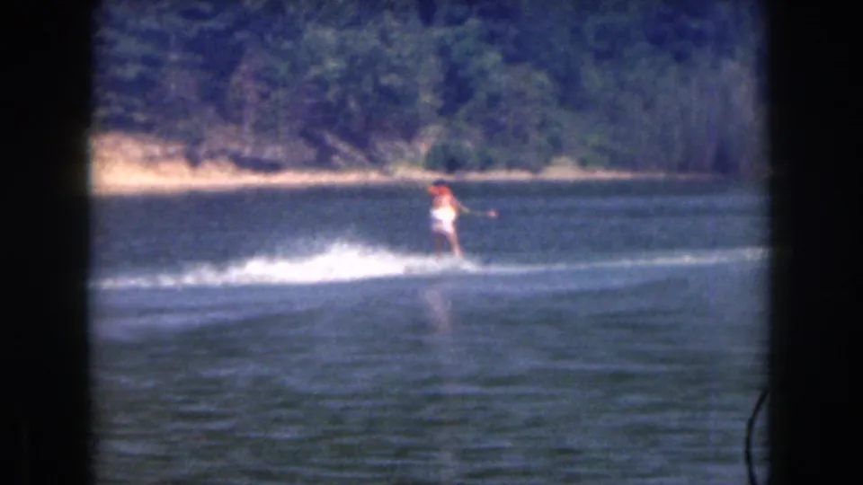 Distant 1961 Michigan lake view of a woman water skiing across open water, with spray, tow line, and wooded shoreline visible.