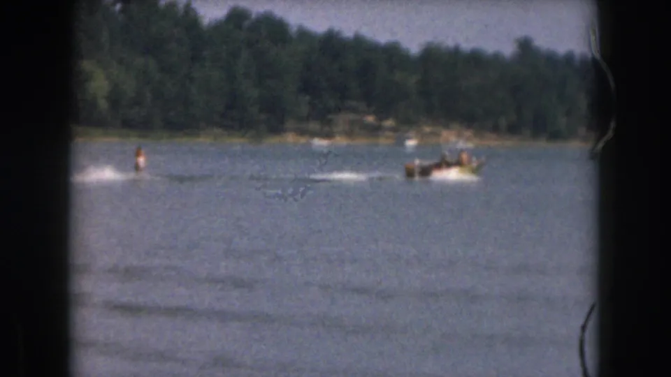 1961 Michigan 8mm frame with a small boat carrying people and a distant skier crossing a tree-lined lake.