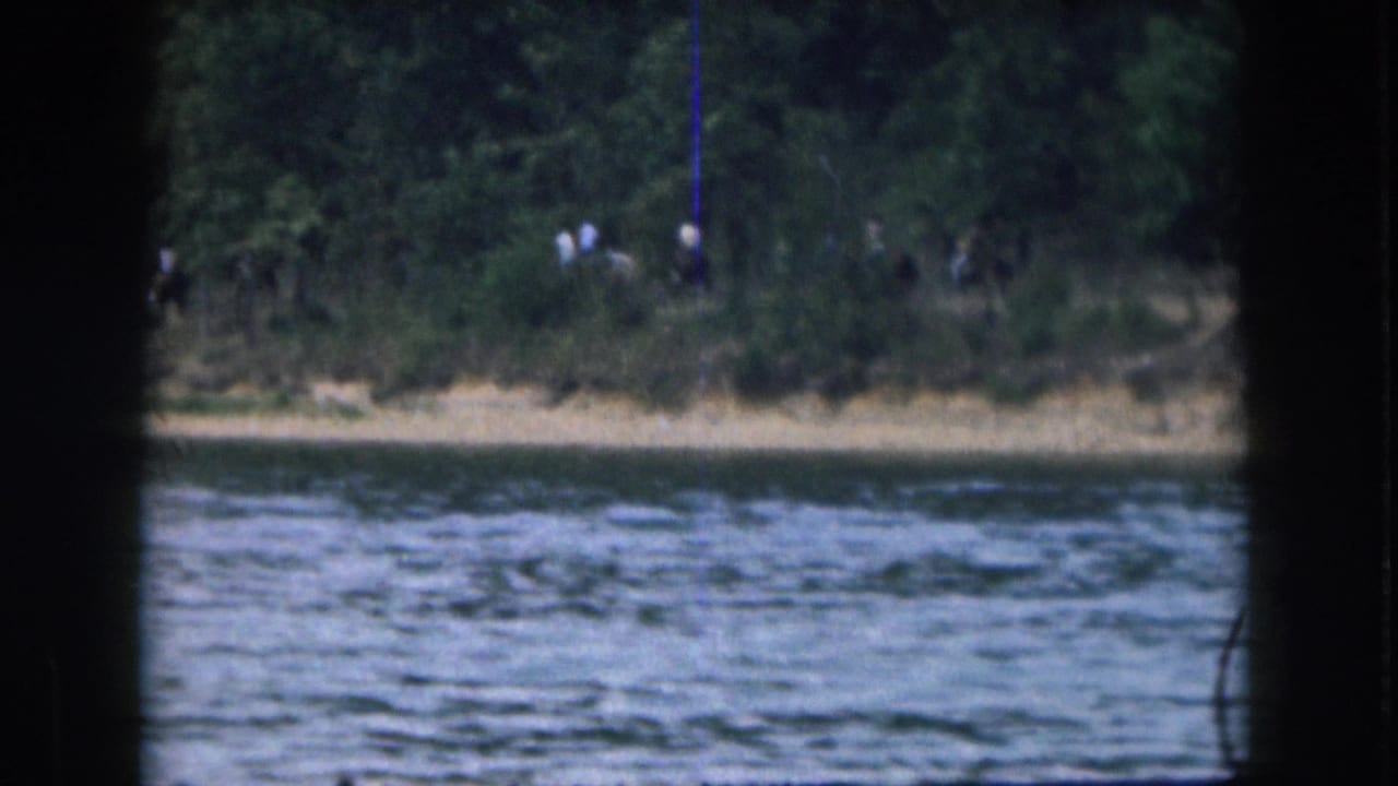 1961 Grand Rapids home-movie frame showing a rippled lake and wooded shoreline, with distant people barely visible near the bank.