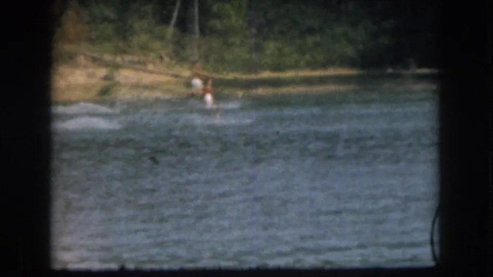 1961 Grand Rapids home-movie frame showing a distant male water skier in white shorts crossing the lake near trees.