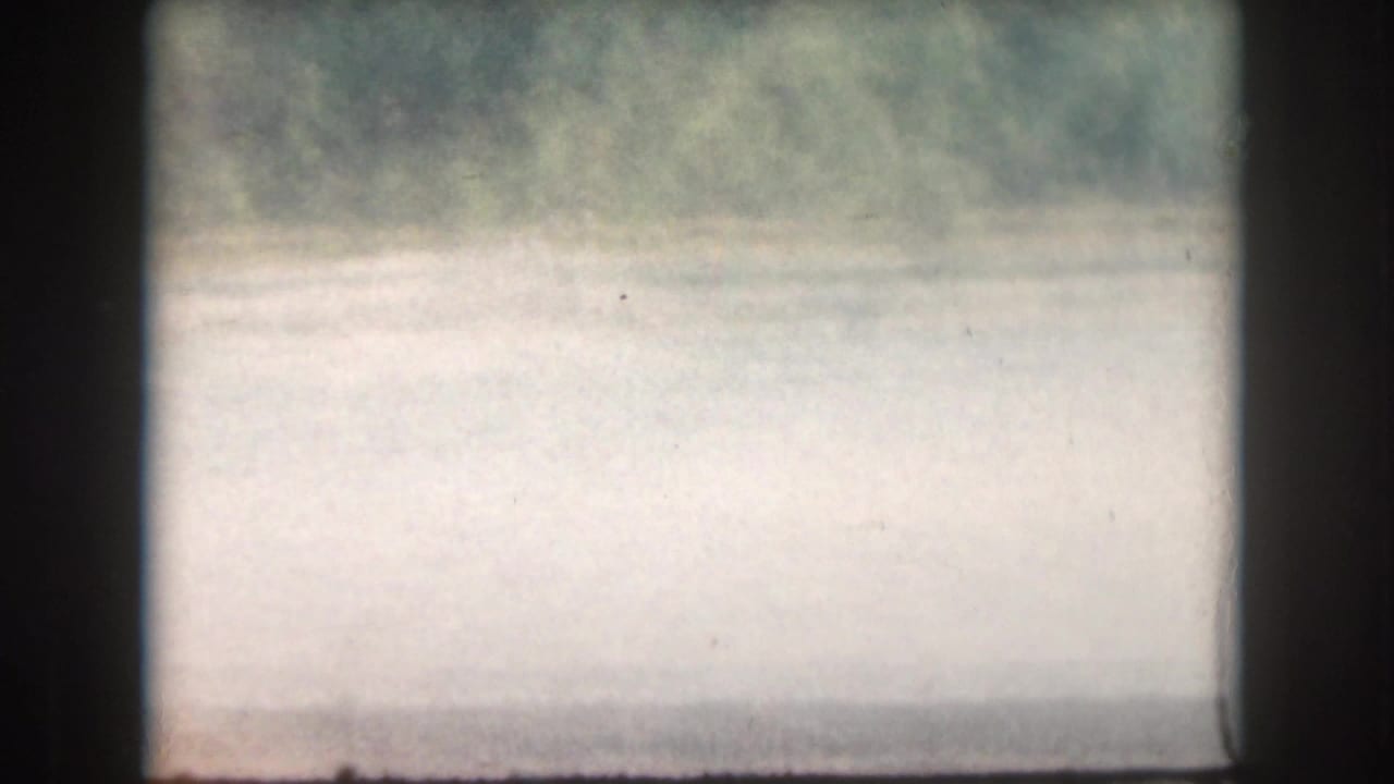 1961 home-movie frame from Grand Rapids, Michigan, showing pale lake water and a blurred shoreline before the skier enters view.