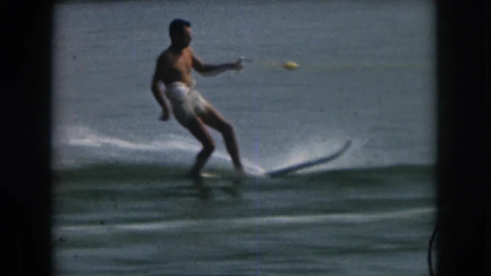 1961 Grand Rapids, Michigan home-movie frame with an adult man in white trunks skiing on a single ski behind an unseen boat.
