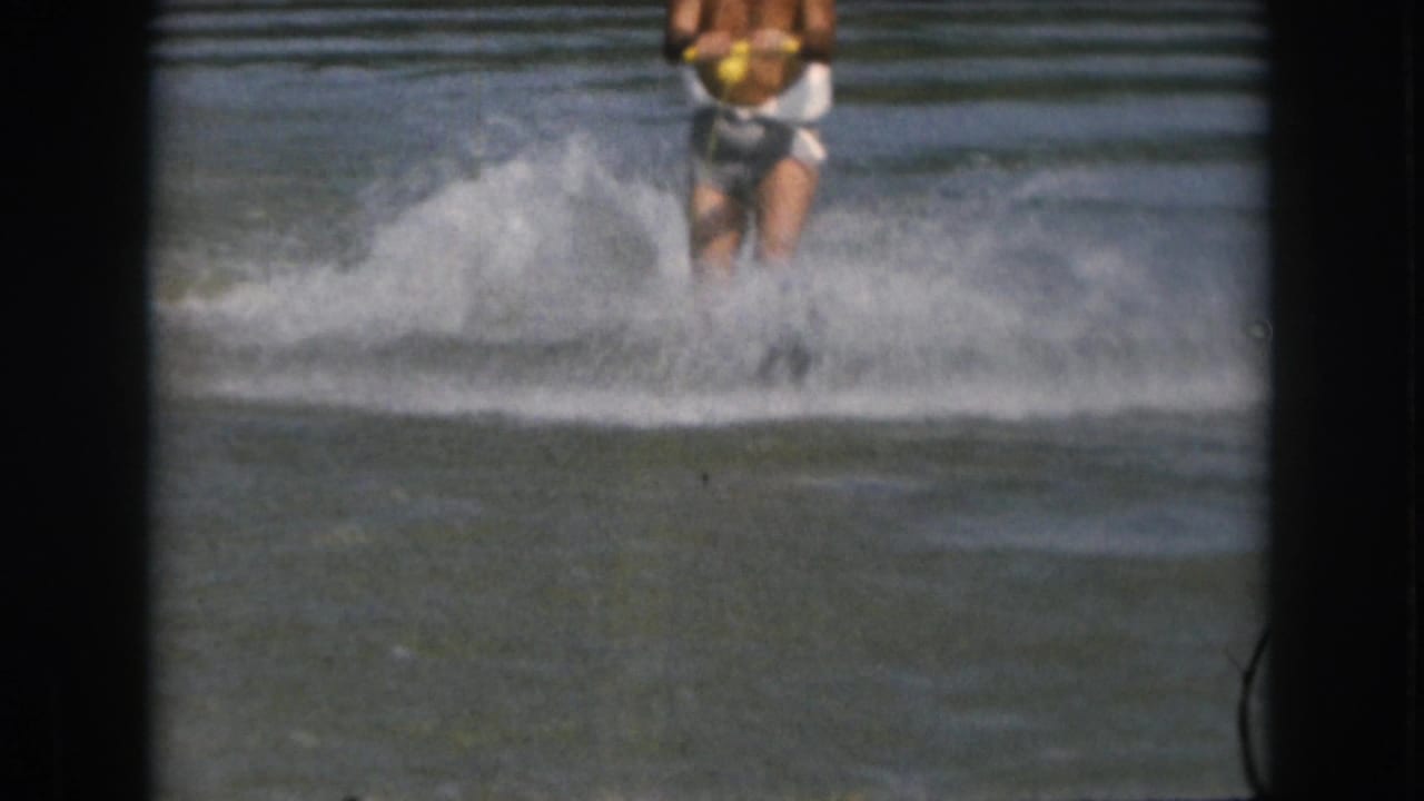1961 Michigan home-movie frame showing a distant male water skier in white trunks holding a yellow tow handle amid lake spray.