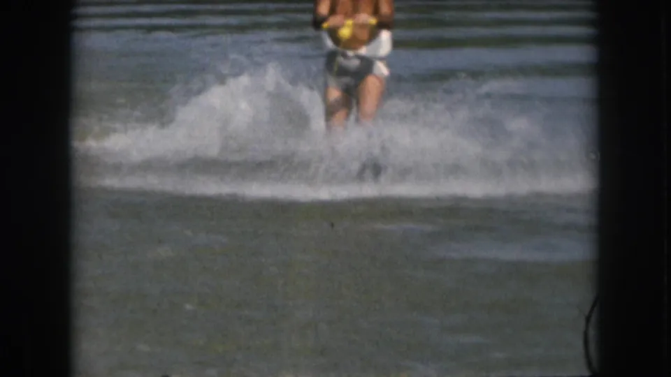 1961 Michigan home-movie frame showing a distant male water skier in white trunks holding a yellow tow handle amid lake spray.