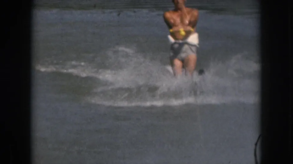1961 Michigan home-movie frame of an adult man water skiing in white trunks and a yellow life vest, seen through soft 8mm film grain.