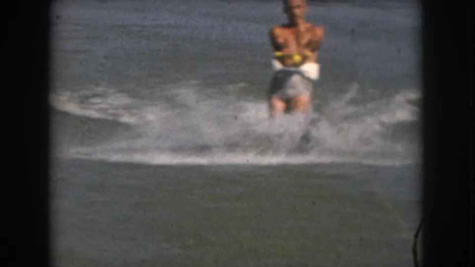 1961 Michigan home-movie frame of an adult man on water skis, centered in choppy water with spray and soft 8mm motion blur.