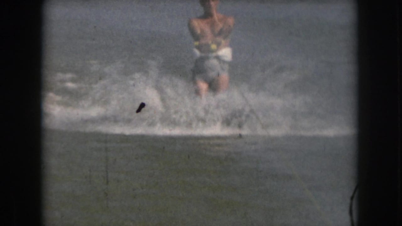 In 1961 Michigan home-movie footage, a shirtless adult man water skis toward the camera while spray rises around him.