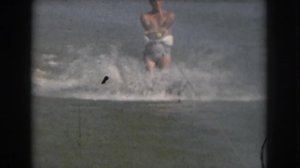 In 1961 Michigan home-movie footage, a shirtless adult man water skis toward the camera while spray rises around him.