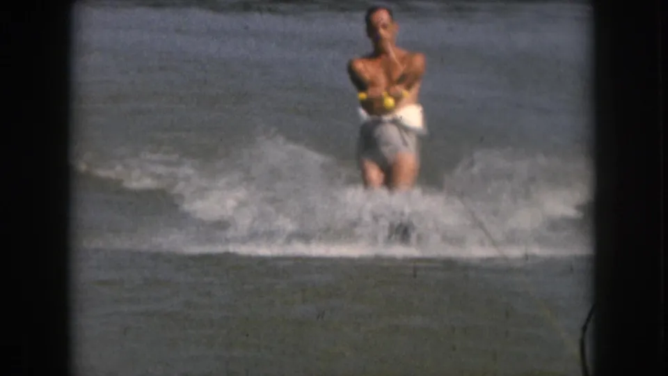 The adult skier grips a yellow tow handle as he moves across the lake in faded 8mm color footage from the Grand Rapids area.