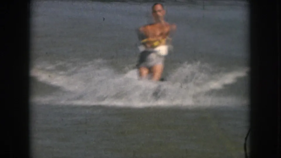 A shirtless adult water skier in light trunks rides toward camera, with soft focus and lake spray emphasizing the home-movie texture.