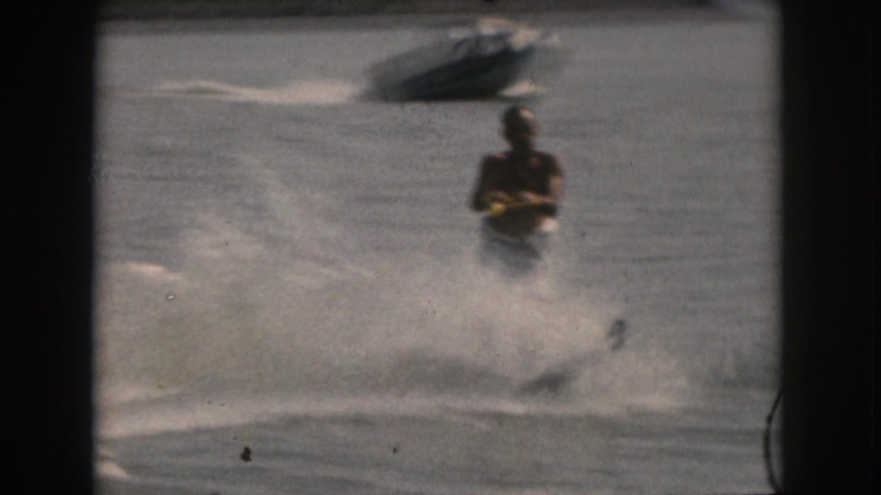 In 1961 Grand Rapids, Michigan area home-movie footage, an adult man water skis across a lake as a towing boat appears in the background and water spray fills the foreground.