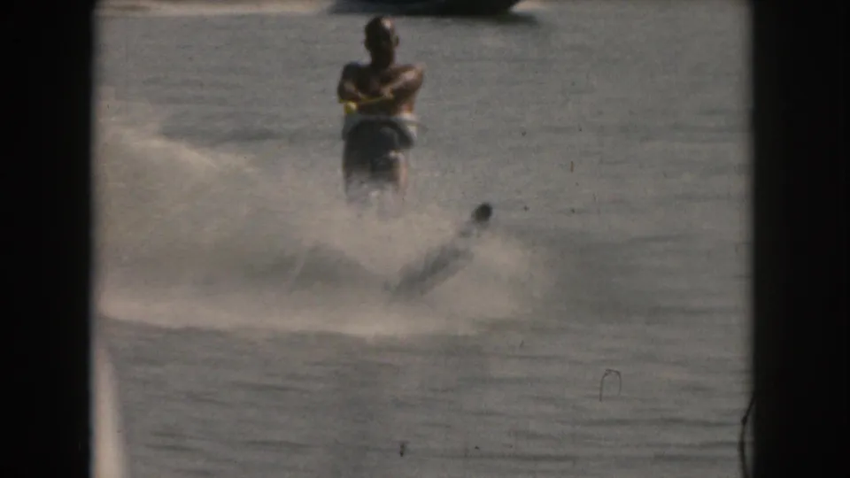 The skier leans into a slalom-style turn on the lake, partly obscured by spray, in soft 1961 8mm home-movie footage from the Grand Rapids, Michigan area.