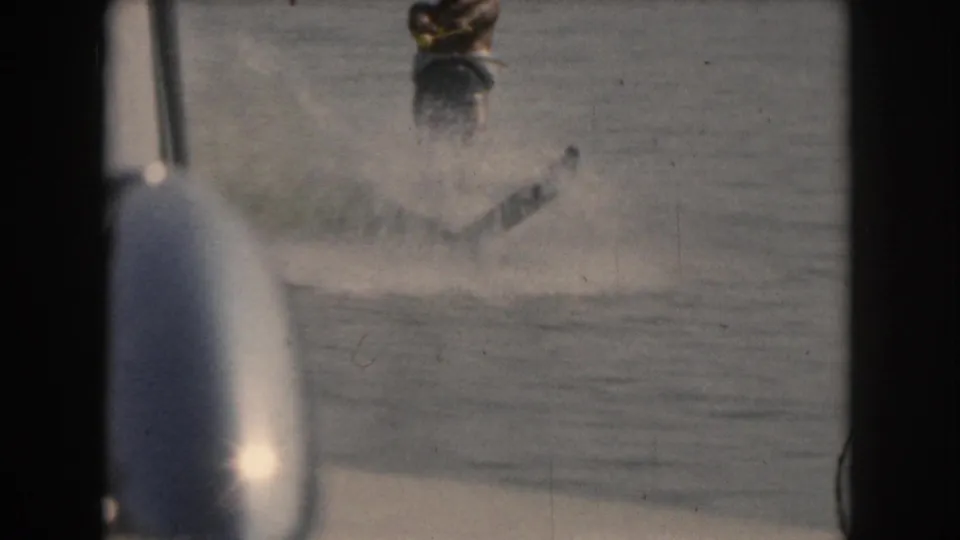 A water skier continues across the lake as a partial foreground boat element enters the frame, with spray and film-edge borders visible in the 1961 Grand Rapids, Michigan area home movie.