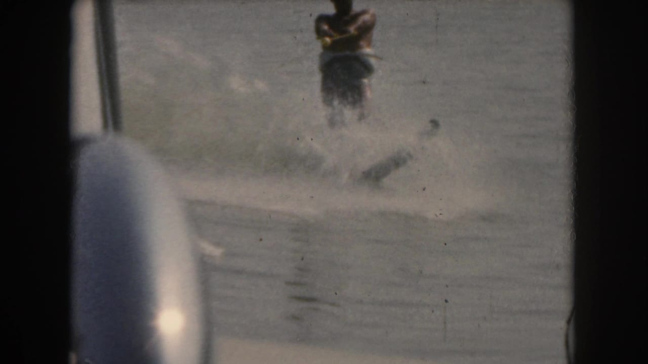 Man water-skiing behind a boat on calm water, partly obscured by boat framing.