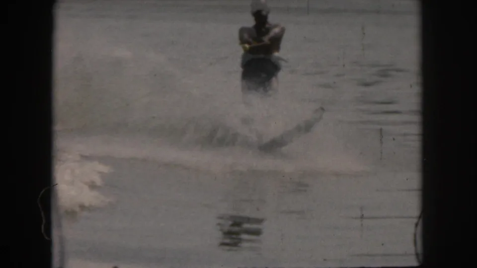 Home-movie frame from the 1961 Grand Rapids clip showing the skier centered behind the boat as water spray spreads across the lake.