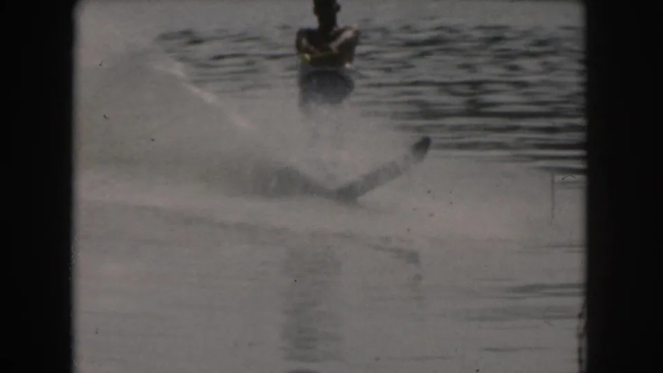 1961 Grand Rapids lake recreation frame with a shirtless water-skier crossing the wake, seen through soft 8mm grain and boat framing.