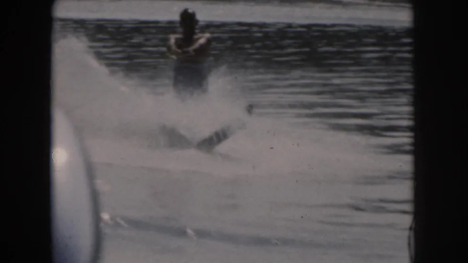 A 1961 Michigan lake scene shows a man water skiing with a fan of spray in grainy 8mm footage.