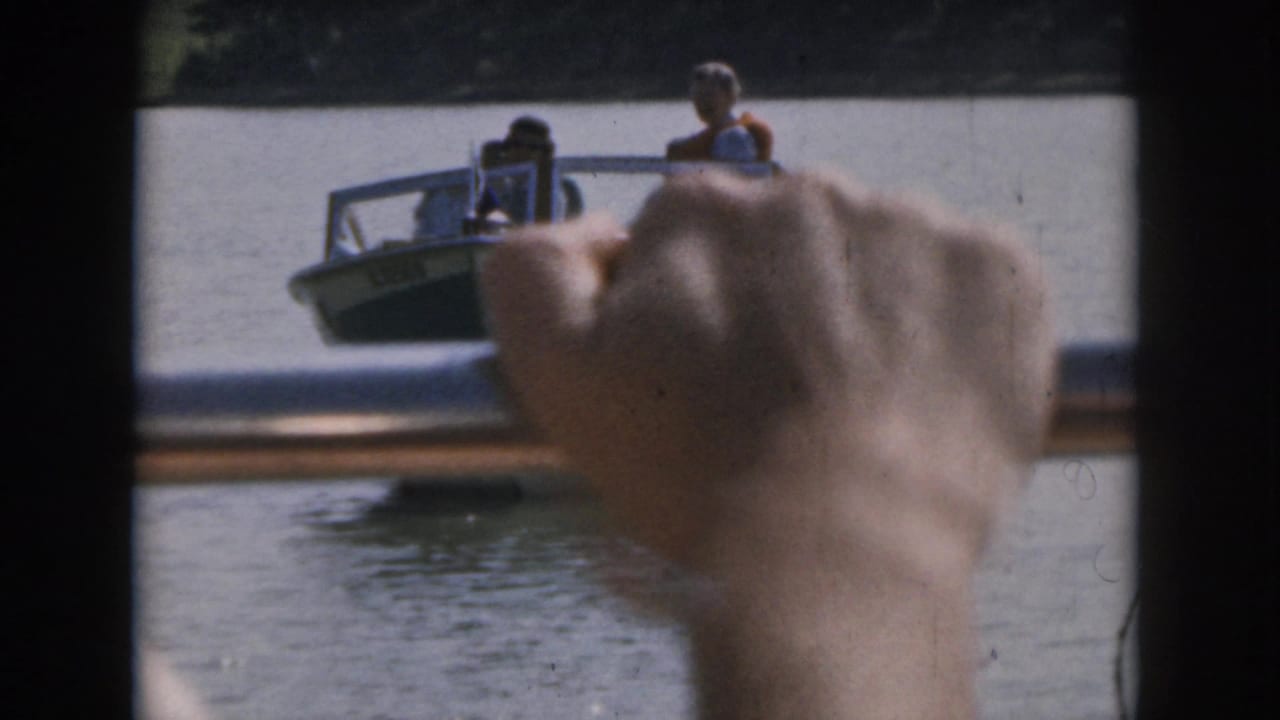 Hand raised in foreground with a small motorboat crossing the lake beyond it.