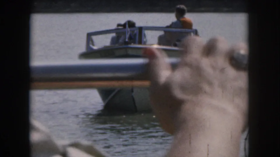 From a moving boat, the camera catches another small motorboat on the lake with a hand, rail, and side mirror partly blocking the view.