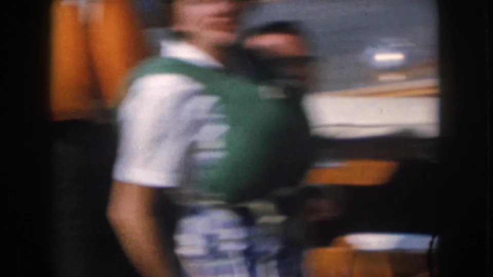 1961 Michigan home-movie frame showing a woman in a green life vest and plaid skirt moving past the camera on a boat.