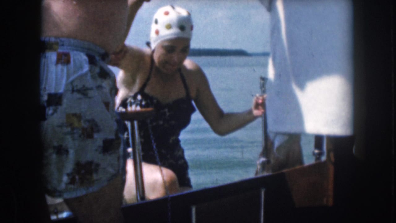 1961 home-movie frame from Michigan shows a woman in a swim cap climbing onto a boat while a man stands nearby.