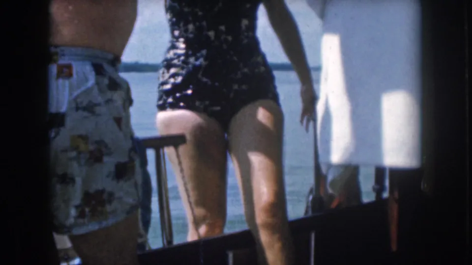 A woman in a dark one-piece swimsuit steps aboard a boat in 1961 Michigan footage, with water and boat hardware behind her.