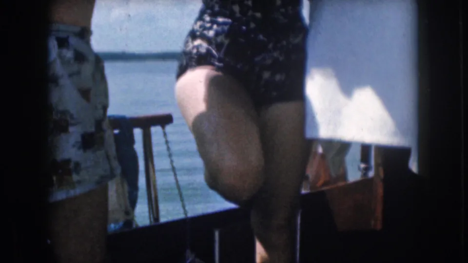 The 8mm frame shows the woman low by the railing after climbing aboard, with a man partly visible and water beyond the boat.