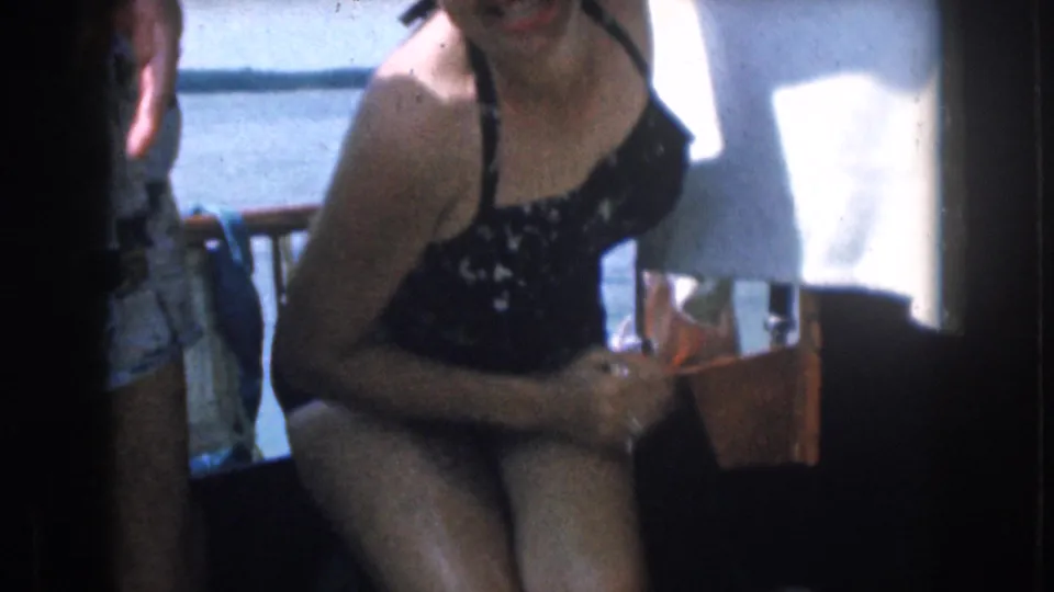 A woman in a dark patterned swimsuit smiles aboard a boat in the 1961 Michigan home-movie footage, with water visible behind her.