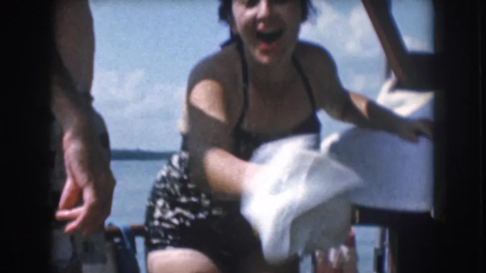 1961 Michigan home-movie frame of a woman laughing and moving a white towel while seated or leaning on a small boat.