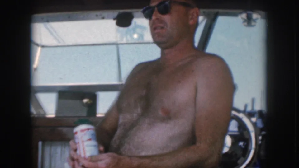 1961 Michigan home-movie frame showing a shirtless man in sunglasses seated on a boat, holding a drink can near the cabin structure.
