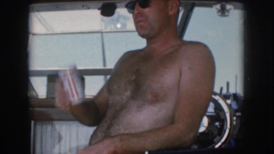 1961 Michigan home-movie frame of a shirtless man in sunglasses raising a drink can while seated inside the boat.