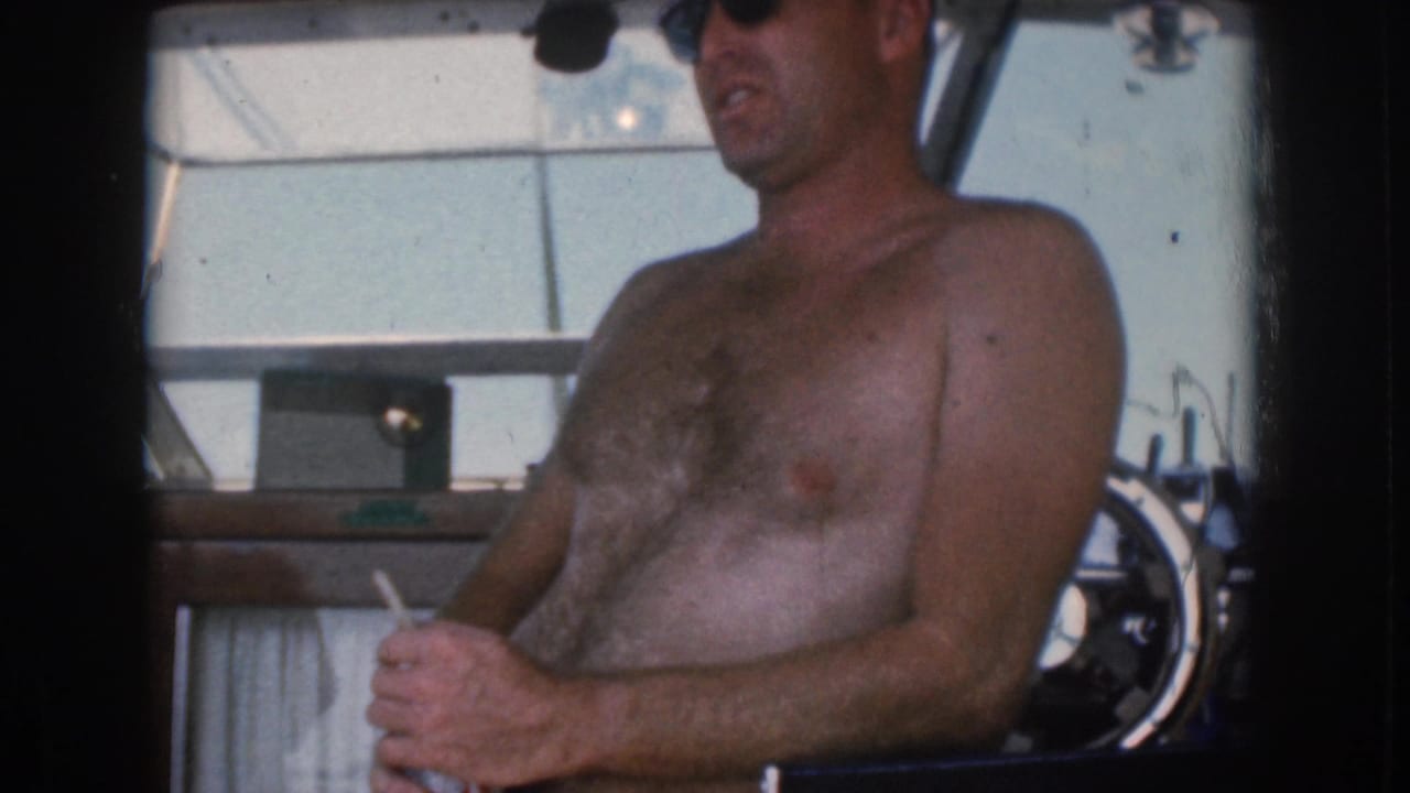 1961 Michigan home-movie frame showing a shirtless man in dark sunglasses at a boat helm with a cigarette in hand.