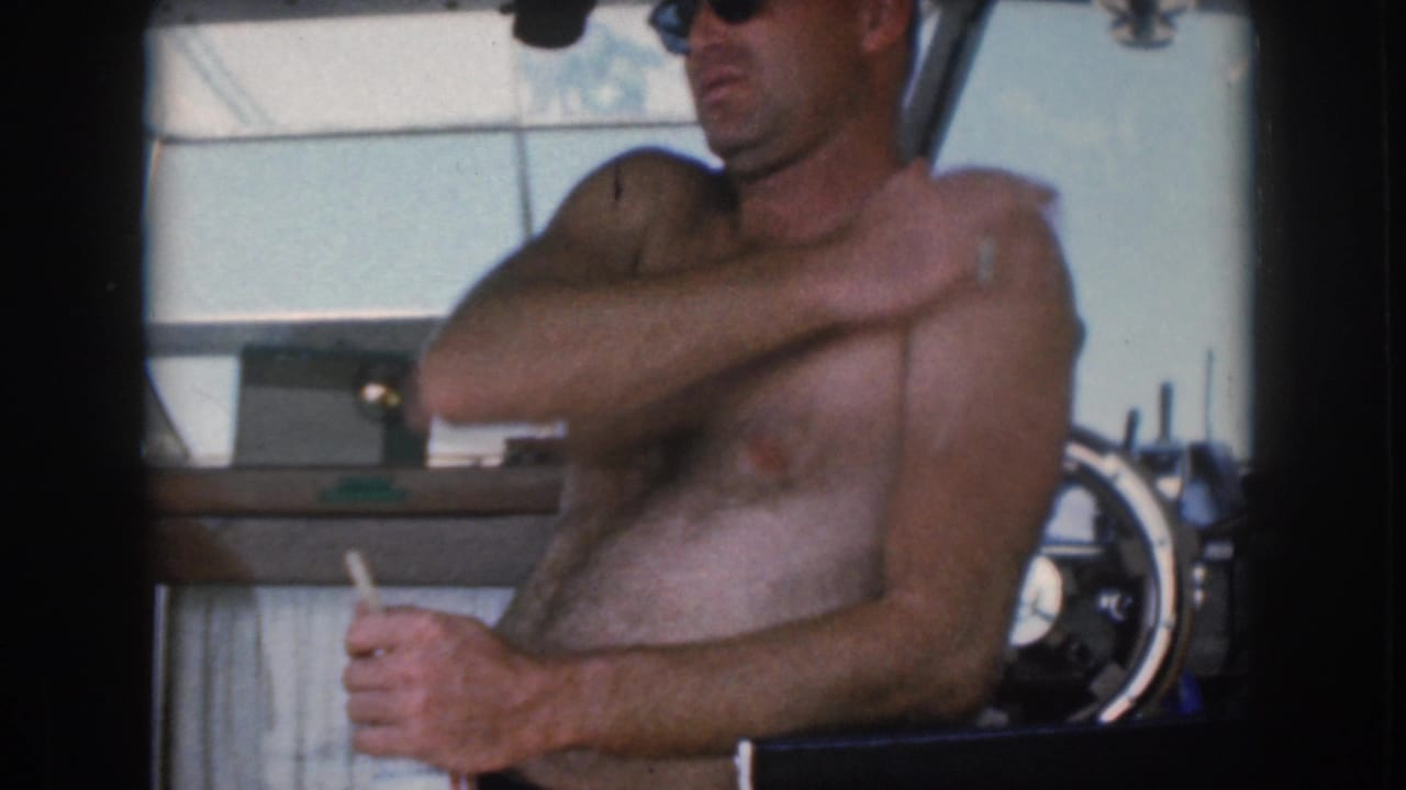Shirtless man in sunglasses at the helm reaches across his shoulder while holding a cigarette.